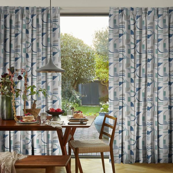 Floor-length pleated curtains with blue-and-teal geometric pattern frame a sliding glass door, pulled aside to reveal a sunlit backyard; wooden dining table with pancakes and pendant light in foreground.
