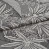 Decorative woven fabric with large stylized white tropical leaves and small flowers, folded into overlapping layers, lying flat in a close-up studio shot with soft, neutral lighting and high-detail texture.