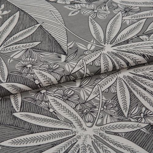 Decorative woven fabric with large stylized white tropical leaves and small flowers, folded into overlapping layers, lying flat in a close-up studio shot with soft, neutral lighting and high-detail texture.