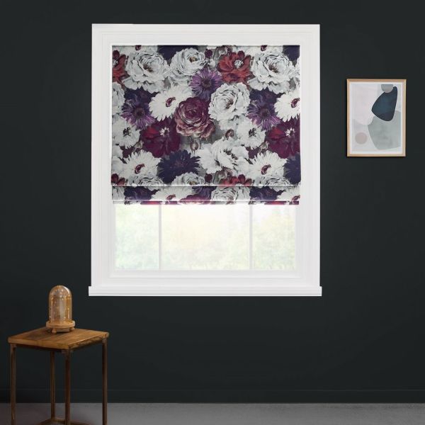 Floral Roman blind, folded and partially lowered, filtering soft daylight; in a white window frame on a dark charcoal wall, beside a wooden side table with glass-domed lamp and abstract print.