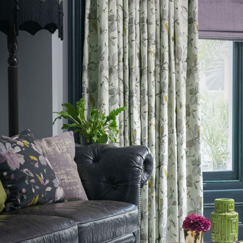 Full-length pleated floral curtains (pale green with grey-yellow blooms), drawn closed, softly filtering daylight; living room setting with black tufted leather sofa, cushions, potted fern, and a teal window frame.