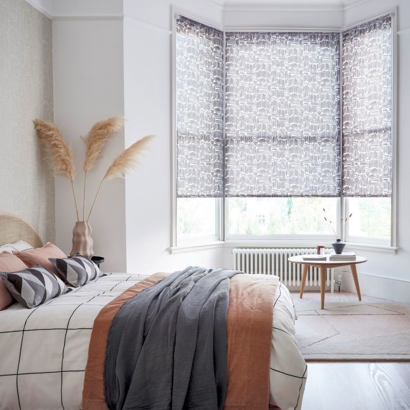 Patterned roller blinds on a three-panel bay window, lowered and filtering soft daylight; bedroom with queen bed, geometric cushions, pampas grass in vase, light wood side table and radiators visible.
