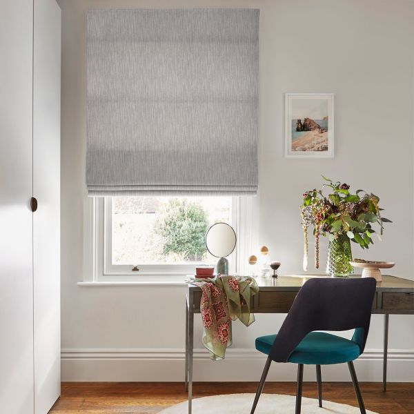 Grey textured Roman blind, lowered and slightly folded at bottom, filtering soft daylight through the window; a desk beside it with chair, mirror, vase of flowers and framed seaside print.