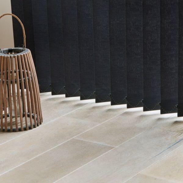 Vertical fabric blinds, dark charcoal, hanging closed with scalloped weighted bottoms; they block most light while small gaps cast narrow beams on a pale tiled floor beside a wicker lantern.