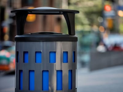 grey metal waste and recycling container with a black plastic canopy lid sitting on a busy city sidewalk