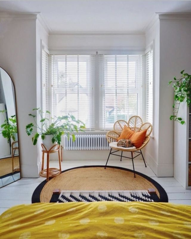 White horizontal slatted blinds covering a three-panel bay window, slightly tilted and filtering bright daylight, in a sunlit bedroom corner with rattan chair, orange cushions, plants and round jute rug.