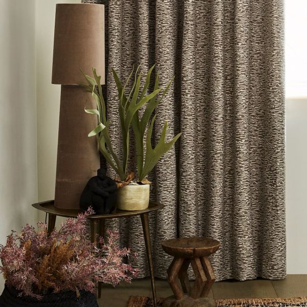 Patterned brown curtains (full-length, heavy fabric) drawn closed, softly filtering light; beside them a side table holds a tall lamp, potted staghorn fern and decorative objects in a warm corner.
