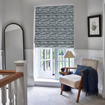 A textured gray-blue Roman blind with horizontal brushstroke pattern, lowered over a sash window, filtering light. In a bright landing: beige armchair with navy throw, lamp, tall mirror, garden view.