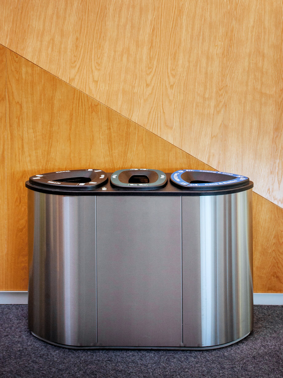 triple stream stainless steel all metal waste and recycling container with hinged lid against a wood panel wall in a university
