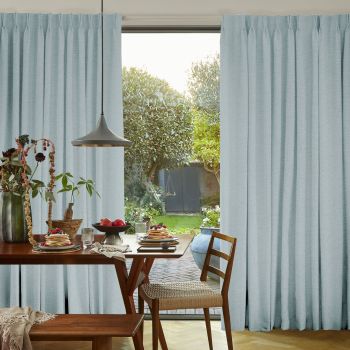 Light-blue pleated curtains, drawn aside to reveal a sliding glass door, filtering daylight into a dining area with a wooden table set for breakfast and a sunlit garden beyond.