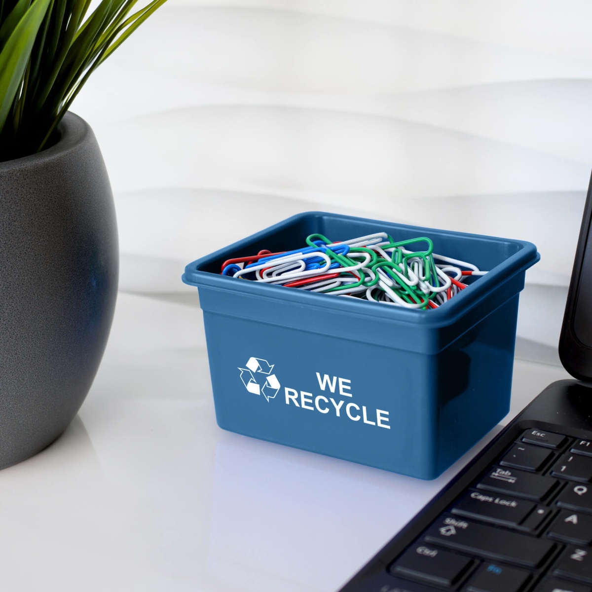 mini blue curbside recycling container with we recycle printed on the side filled with paper clips sitting on an office desk
