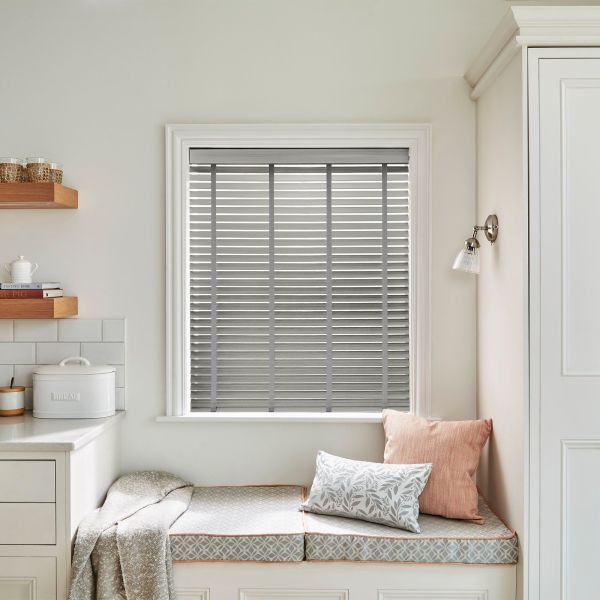 Grey horizontal blinds with vertical ladder tapes covering the window, closed and filtering soft daylight; cozy kitchen nook with built-in cushioned bench, pillows, wall sconce, countertop and bread tin.
BREAD