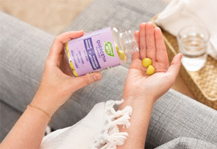 A hand pouring out two gummies from a bottle of Nature's Way Quercetin with Zinc gummies with the arm of a couch and a glass of water on a tray in the background.