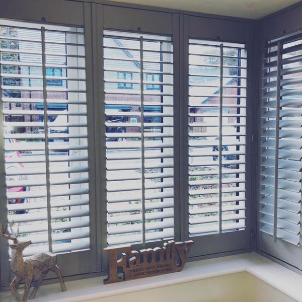 White wooden plantation shutters cover a three-panel bay window, slats tilted open to filter bright daylight and show a suburban street; windowsill holds a wooden "Family" sign and deer ornament.

Transcription:
Family
WHERE LIFE BEGINS & LOVE NEVER ENDS