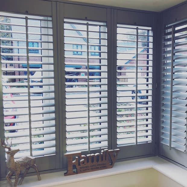 White wooden plantation shutters cover a three-panel bay window, slats tilted open to filter bright daylight and show a suburban street; windowsill holds a wooden "Family" sign and deer ornament.

Transcription:
Family
WHERE LIFE BEGINS & LOVE NEVER ENDS