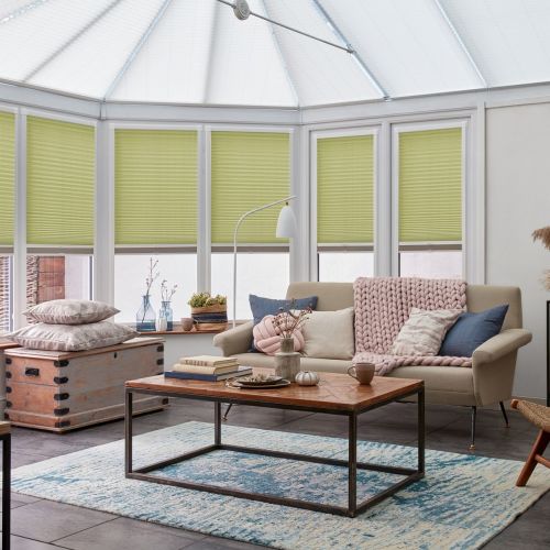 Lime-green pleated cellular blinds, lowered across conservatory windows and filtering daylight; set in white frames beneath a translucent roof, overlooking a cozy living area with sofa, coffee table and rug.