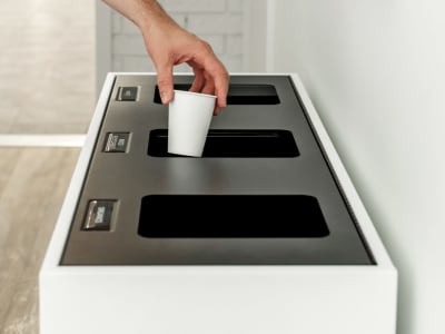 white metal recycling and trash bin with black hinged lid showing a person putting a paper cup into the recycling stream