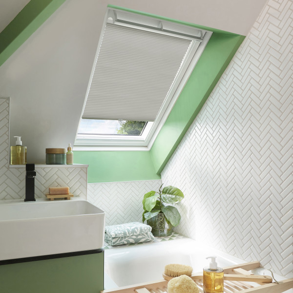 A white pleated skylight blind, mounted inside a sloped roof window, lowered to filter daylight; bathroom with mint-green trim, white herringbone tiles, sink, tub, plant and toiletries.