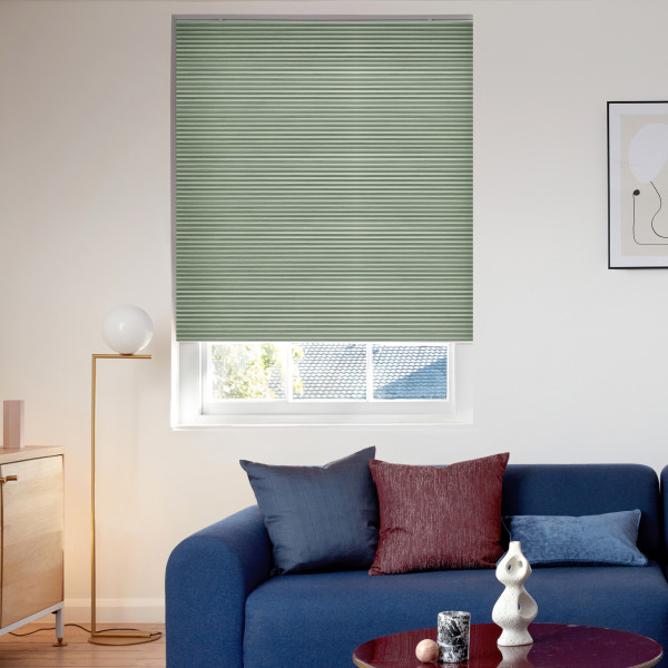 Sage-green pleated cellular blind, lowered with thin horizontal ribs and bottom edge slightly above the sill, blocking light; in a sunlit living room with a blue sofa, cushions, and a floor lamp.