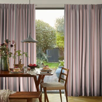 Floor-to-ceiling pleated striped curtains (gray and pink), parted centrally to reveal a glass door, filtering daylight into a dining room with wooden table, lamp, chair and sunlit garden beyond.