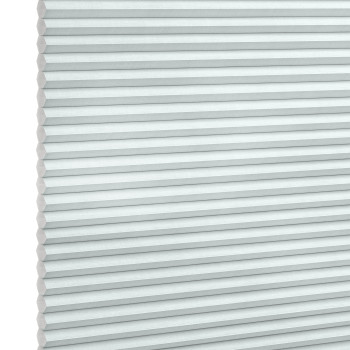 Pleated honeycomb blind, pale gray-blue with hexagonal cells along the left edge, fully lowered and closed, softly filtering daylight, set against a bright white background in a neutral-lit interior.
