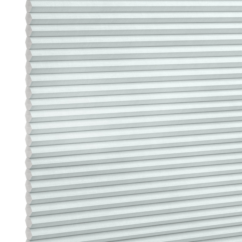 Pleated honeycomb blind, pale gray-blue with hexagonal cells along the left edge, fully lowered and closed, softly filtering daylight, set against a bright white background in a neutral-lit interior.