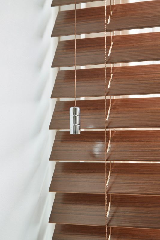 Wooden horizontal venetian blinds with brown wood-grain slats and pull cords bearing a chrome weight; partially closed, slightly angled to filter light in a close-up against a white wall.