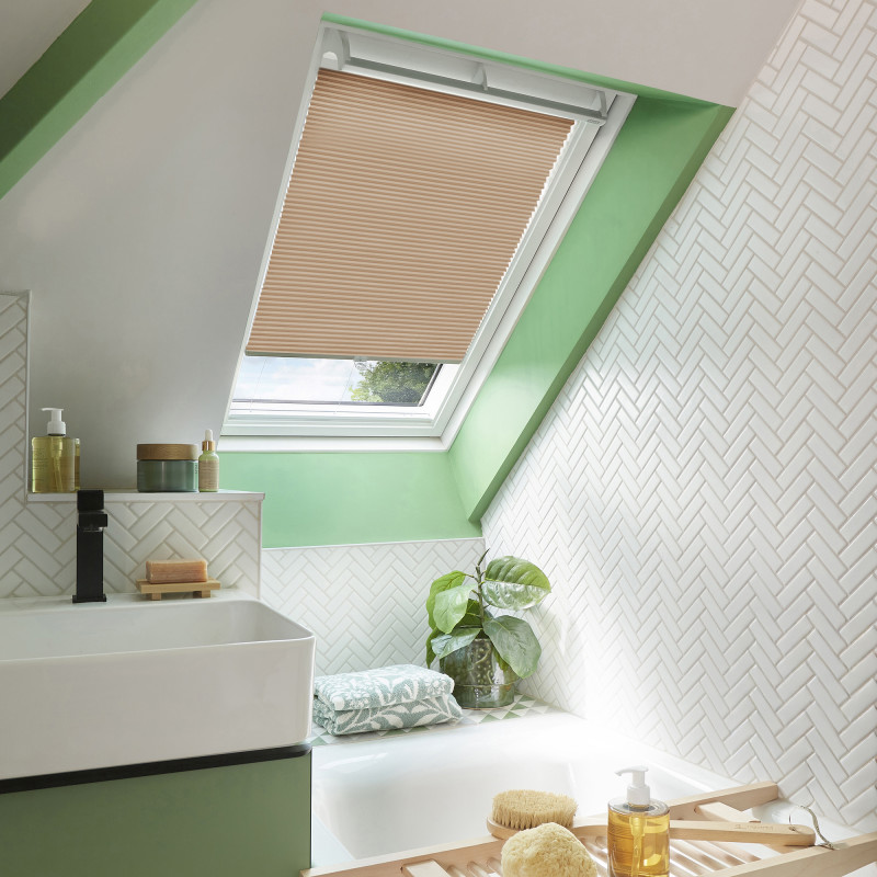 Beige pleated skylight blind, lowered halfway and filtering light, set within a mint-green dormer; surrounding attic bathroom shows white herringbone tiles, sink, bathtub, plant, and bath accessories.