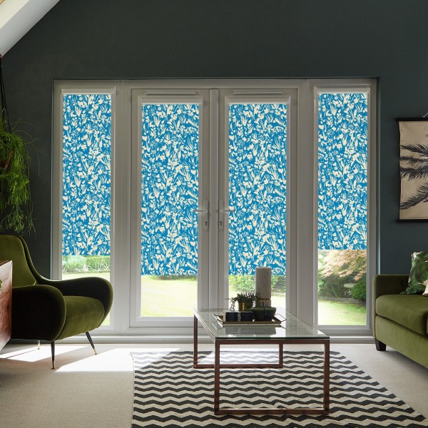 Blue floral roller blinds cover four tall glass doors and sidelights, filtering daylight in a living room with green armchairs, glass coffee table, zigzag rug, and dark-painted walls.