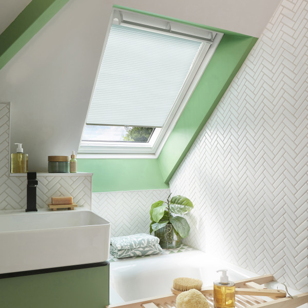 White pleated skylight blind, partially lowered across a slanted roof window, filtering soft daylight; bathroom with green-painted trim, white herringbone tiles, basin, bathtub ledge with plant and toiletries.