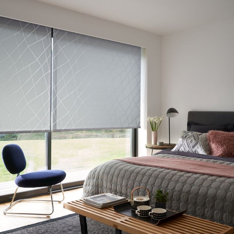 Light-grey patterned roller blinds, lowered across a wide sliding window, filtering daylight. Modern bedroom context: quilted grey bed, pink throw and pillows, bedside lamp, navy lounge chair, lawn visible outside.