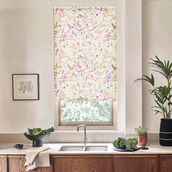 Floral-patterned Roman blind covers a kitchen window; it hangs above a sink and faucet, amid a countertop with bowls of vegetables, potted plants, and a framed sketch.