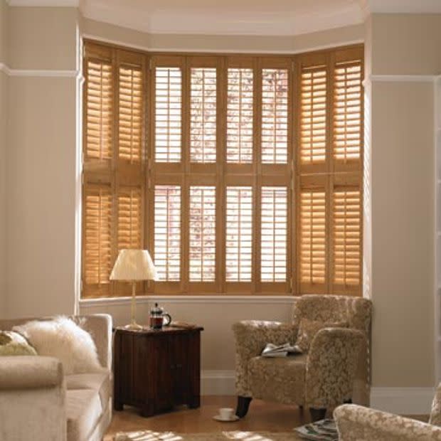 Wooden plantation shutters span a multi-panel bay window, slats partially angled to filter warm daylight; living room setting with upholstered chairs, side table, lamp and soft neutral decor.