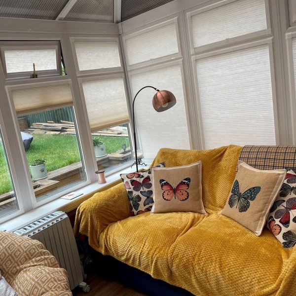 White pleated cellular blinds across a conservatory bay window, mostly lowered and diffusing daylight, two lower panes partially raised revealing lawn; cozy sunroom with mustard throw and butterfly cushions.