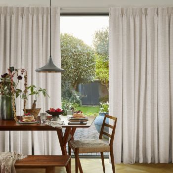 Floor-length beige pleated curtains drawn mostly closed with a central gap; filtering soft daylight and revealing a sunlit garden, foreground dining table set for breakfast.