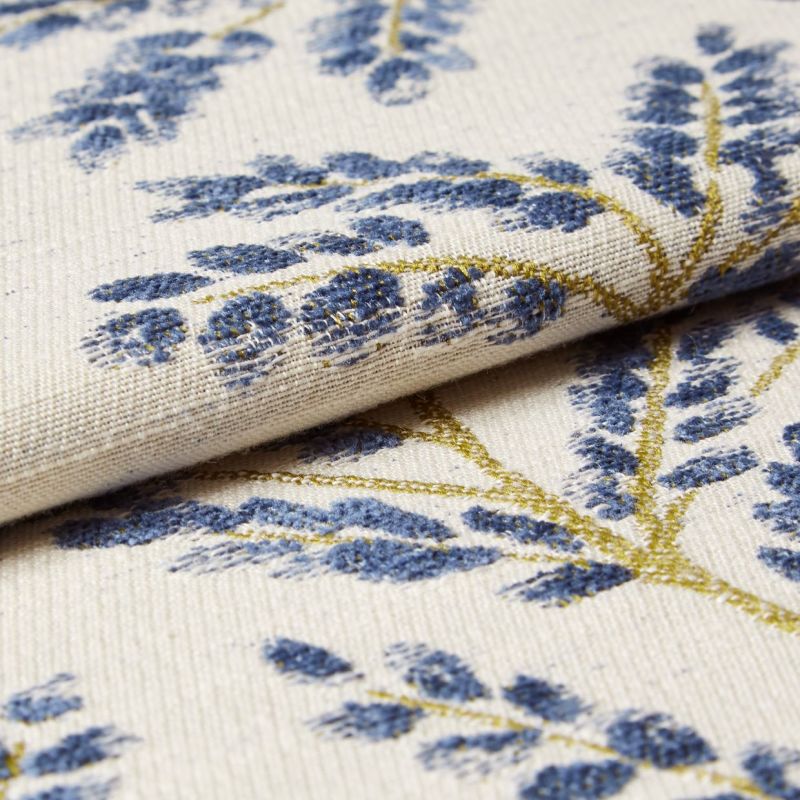 Cream woven curtain fabric with raised blue floral motifs and gold-thread stems â€” folded and layered, showing textured weave â€” close-up studio-lit textile sample with soft, neutral background.
