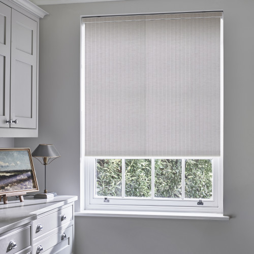 Striped pleated roller blind, lowered nearly to the sill with thin vertical pinstripes; blocking and softly diffusing daylight over a pale-grey room with white cabinetry, lamp, and leafy outside view.
