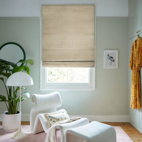 Beige patterned Roman blind, lowered over the window with a small bottom gap, filtering soft daylight; a pastel-green sitting nook with a white sculptural lounge chair, plant, floor lamp, and wooden floor.