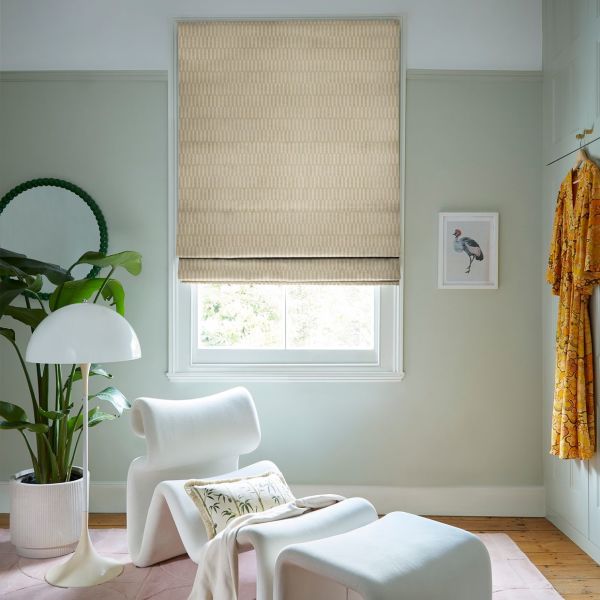 Beige patterned Roman blind, lowered over the window with a small bottom gap, filtering soft daylight; a pastel-green sitting nook with a white sculptural lounge chair, plant, floor lamp, and wooden floor.