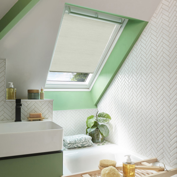 Cream pleated skylight blind, lowered over a sloped roof window, filtering soft daylight into a bright attic bathroom with green trim, white herringbone tiles, sink, tub and plant.