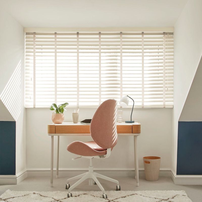 White horizontal Venetian blinds across a wide window, slats tilted with fabric tapes; filtering bright daylight and casting striped shadows; attic home-office niche with wooden desk, pink swivel chair, lamp.