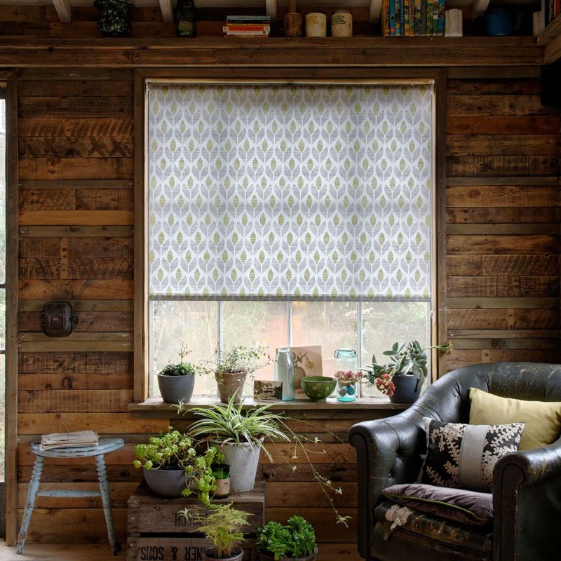 Patterned roller blind (leaf motif) lowered over the window, filtering soft daylight; rustic wood‑paneled room with potted plants on the sill, a worn leather armchair and muted natural lighting.