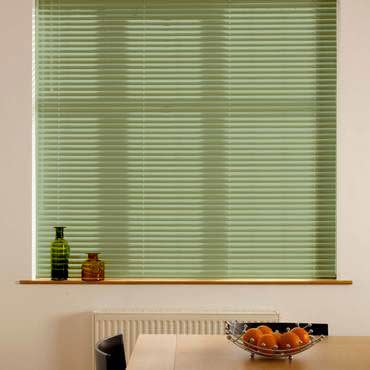 Pale green horizontal blinds, lowered and closed, diffusing daylight and obscuring the view; below them a sill with two bottles, a radiator, and a table with a bowl of oranges.