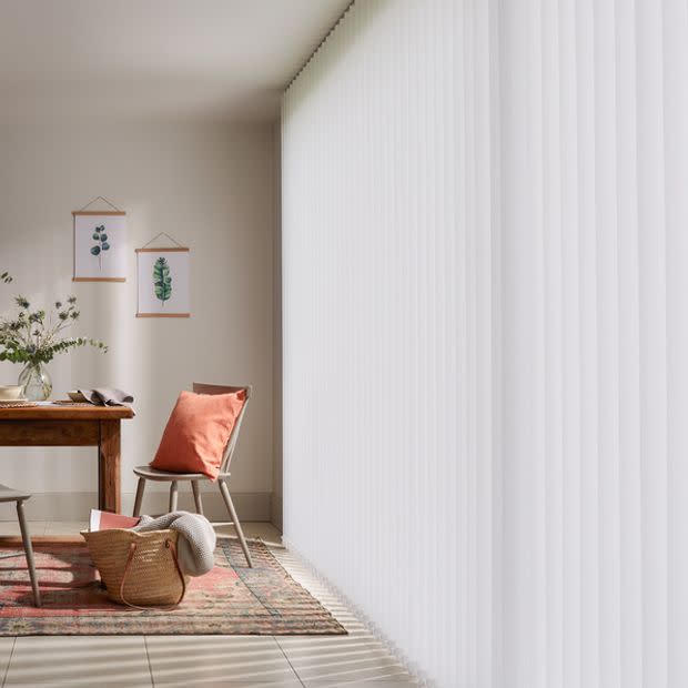 White vertical blinds covering a floor-to-ceiling window, closed and softly filtering sunlight; dining nook with wooden table, chair with terracotta cushion, patterned rug, wicker basket, botanical prints, bright daylight.