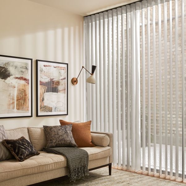Vertical white fabric blinds covering a floor-to-ceiling sliding window, partially closed and filtering soft daylight into striped shadows; living room with beige sofa, textured cushions, abstract wall art, wall lamp.