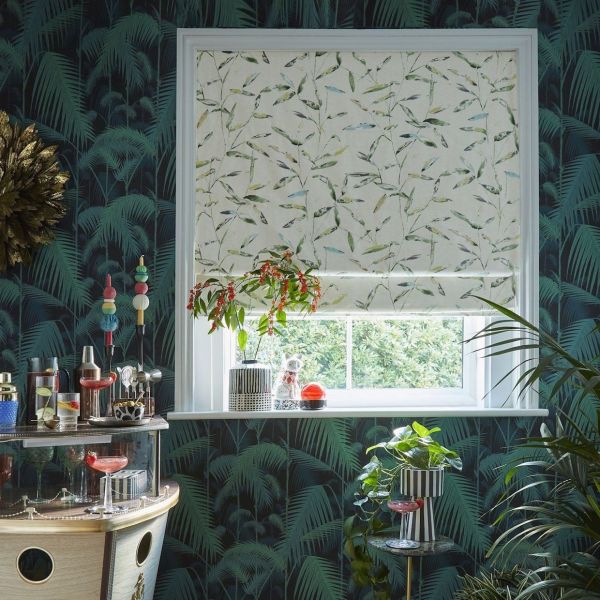 A leaf-printed Roman blind, mostly lowered across a white-framed window, filtering daylight; surrounded by dark palm wallpaper, houseplants, and a small bar cart.