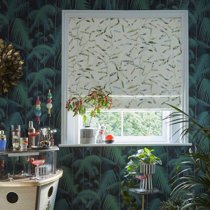 A leaf-printed Roman blind, mostly lowered across a white-framed window, filtering daylight; surrounded by dark palm wallpaper, houseplants, and a small bar cart.