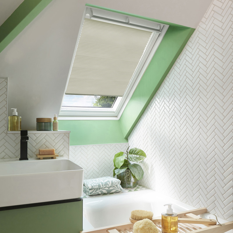 Cream pleated skylight blind, partially lowered and filtering daylight; set in a green-framed roof window above a white-tiled attic bathroom with sink, bathtub ledge, plant, towels and bath accessories.