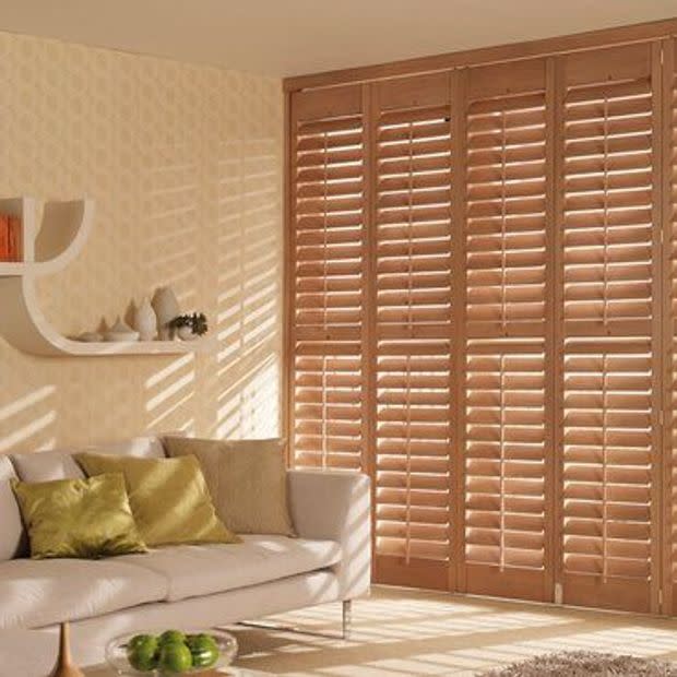Floor-to-ceiling wooden plantation shutters covering a window/door, slatted and partially closed, angling sunlight through; in a warm, sunlit living room with beige sofa, green cushions and shelf.