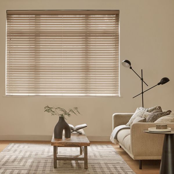 Beige horizontal slatted blinds covering a large window, tilted closed and softly filtering daylight; a beige sofa, wooden coffee table with sculptural vase and open book, black floor lamp, rug.